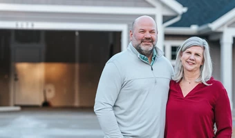 Two adults standing together in front of a suburban home with an open garage. The pair stands close side by side in the driveway, with the house exterior and garage interior softly blurred in the background. 