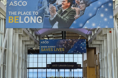 Conference venue hallway with large ASCO banners overhead reading “The Place to Belong” and “Where Science Saves Lives.”