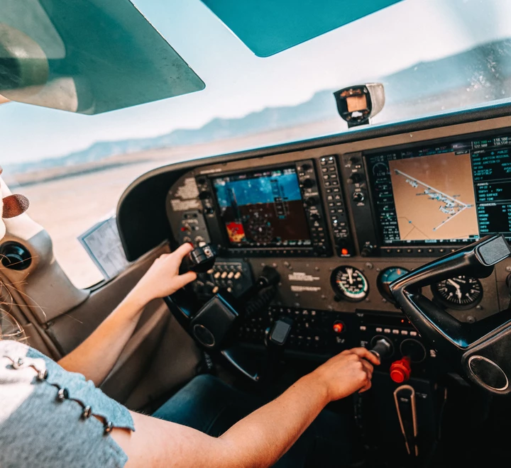 Young adult female pilot in cockpit looking out the window with hands on the controls. Composition is taken inside the airplane looking out at the windshield; pilot is in lower left-side of frame. The small single engine airplane is taxiing on the runway. 