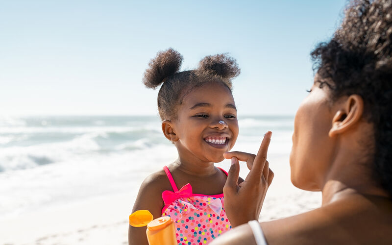 Young African American mother applying protective sunscreen on daughter's nose at beach.