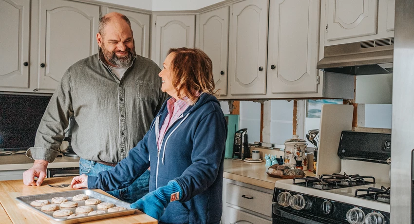 Two people in a home kitchen standing beside a baking sheet of freshly baked cookies on a wooden countertop near a gas stove.