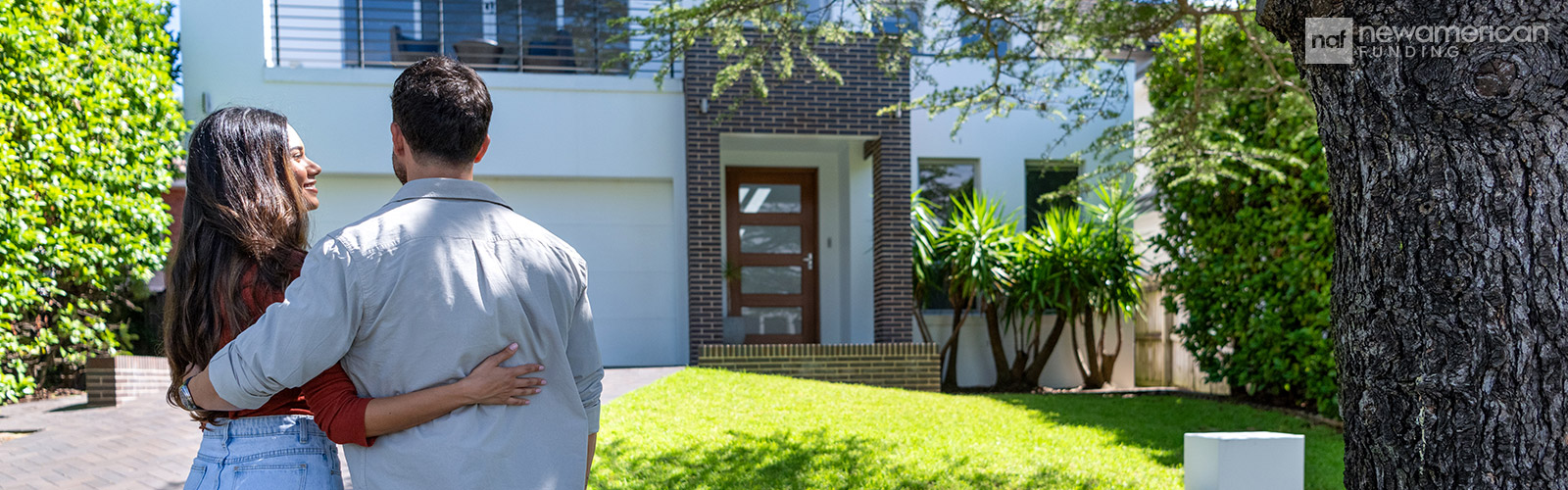 A man and a woman stand smiling with their arms around each other looking at a house