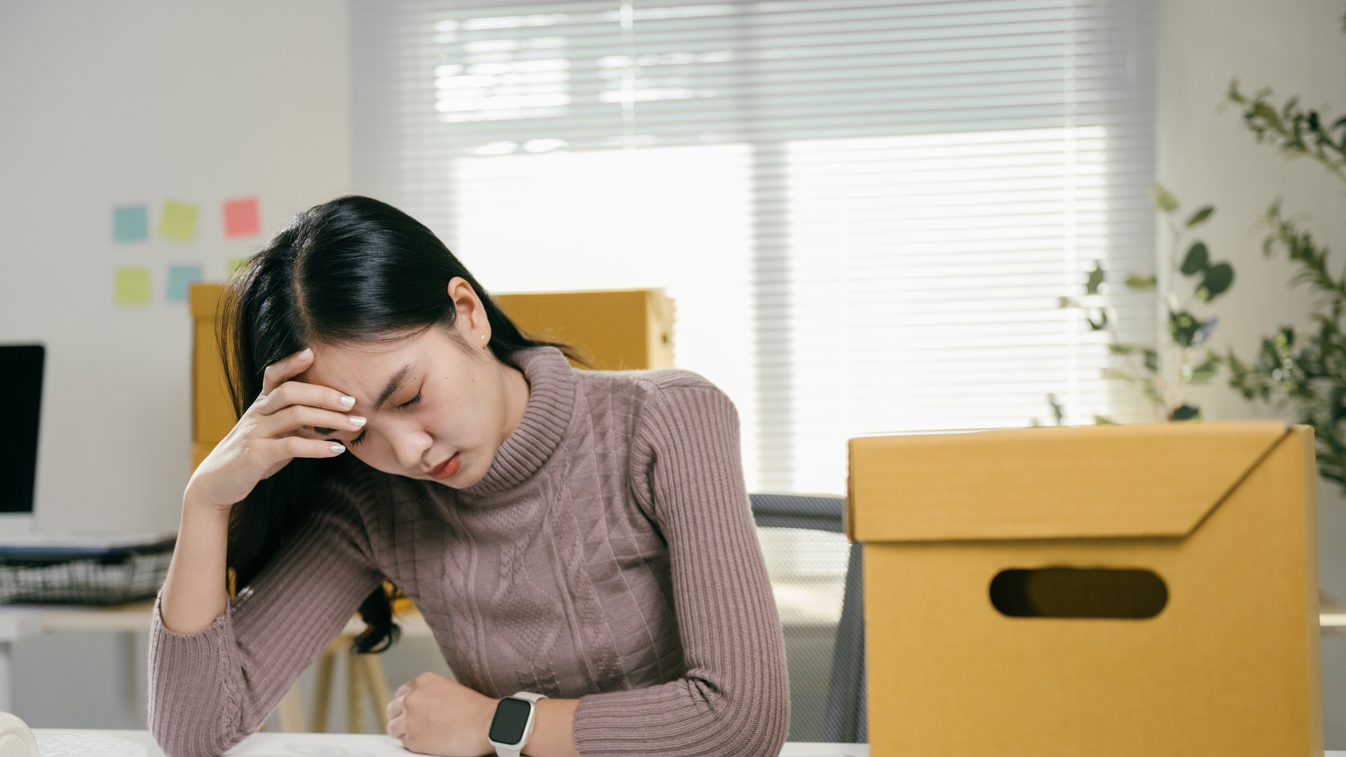 Young asian businesswoman feeling stressed and having a headache while packing her belongings in cardboard boxes, getting ready to leave the office after losing her job or resigning