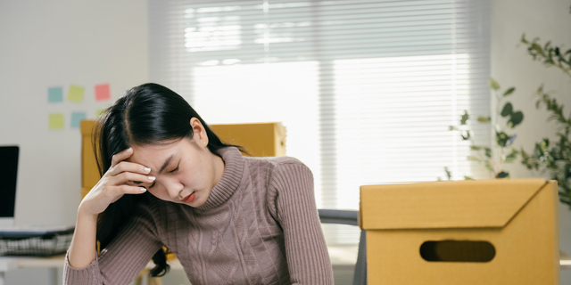 Young asian businesswoman feeling stressed and having a headache while packing her belongings in cardboard boxes, getting ready to leave the office after losing her job or resigning