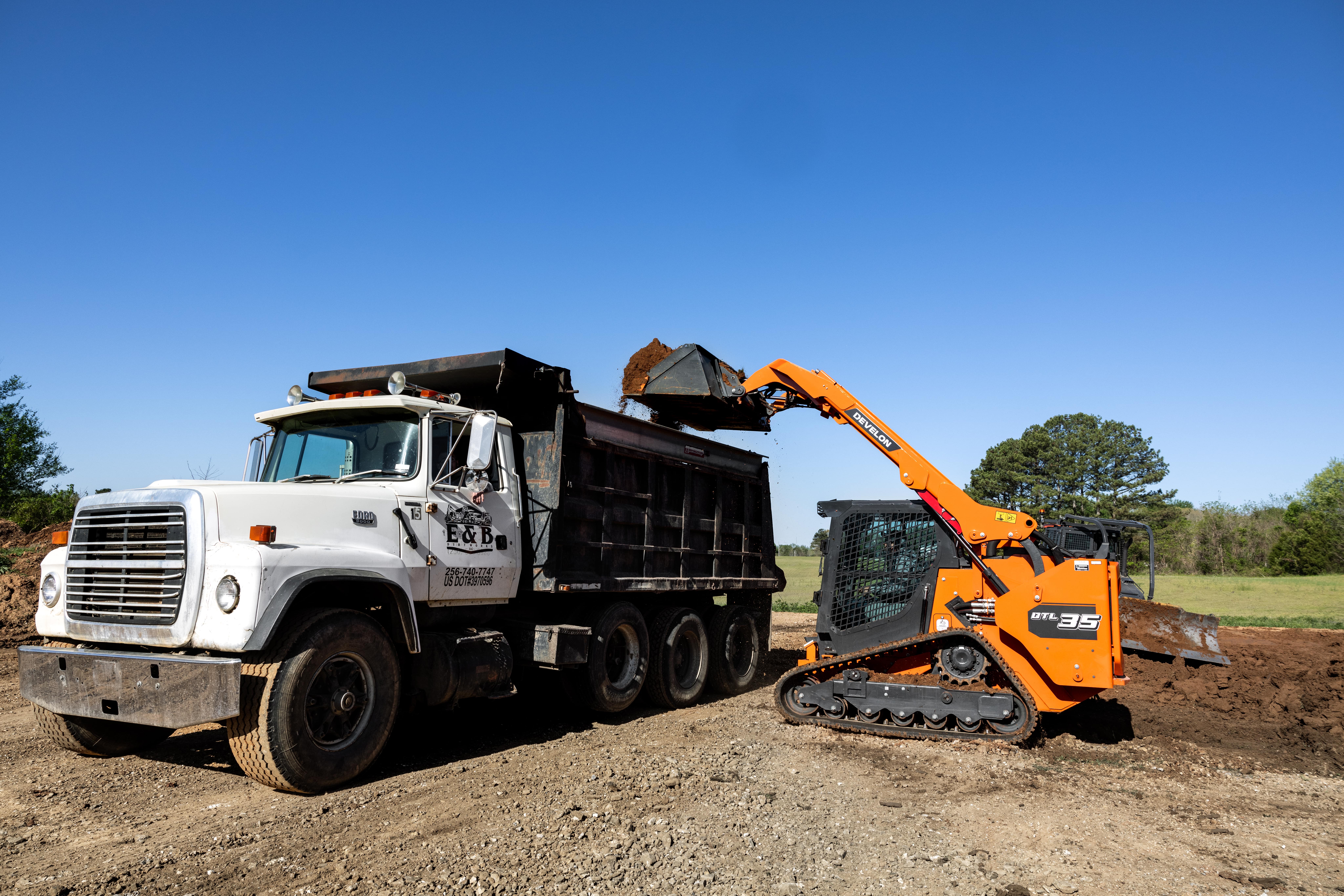 A DEVELON DTL35 compact track loader lifting a bucket filled with dirt into a high-sided dump truck.