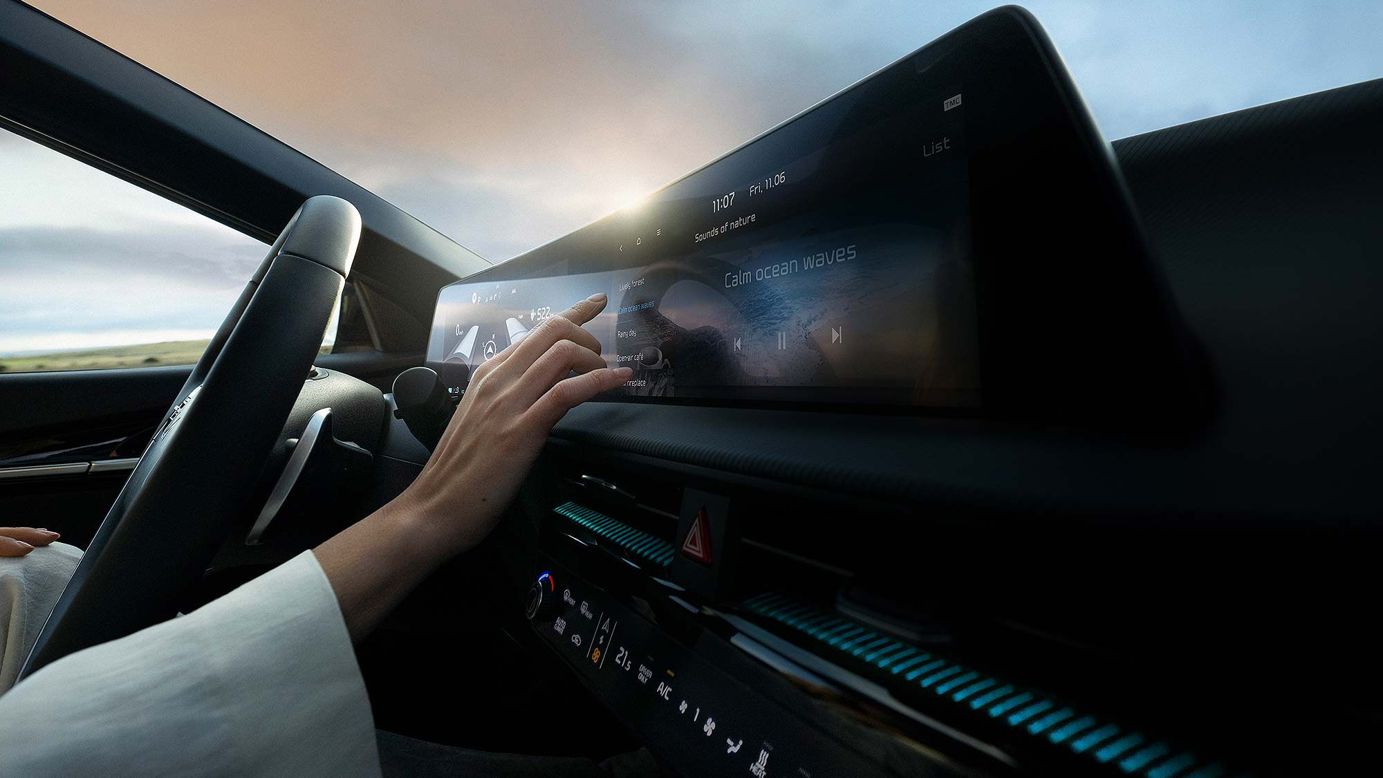 A hand reaches toward a wide in-car touchscreen displaying a “Sounds of nature” interface with options like “Calm ocean waves,” while the car is driven through a scenic landscape at sunset.
