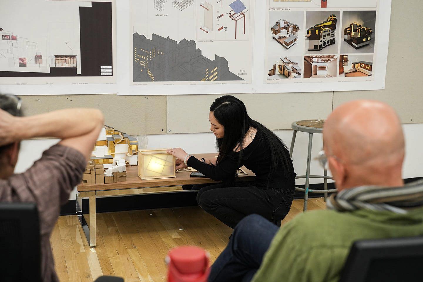 Student Daisy Chen kneels in front of her shadow puppet theater model, demonstrating its internal lighting scheme to an audience during a presentation.
