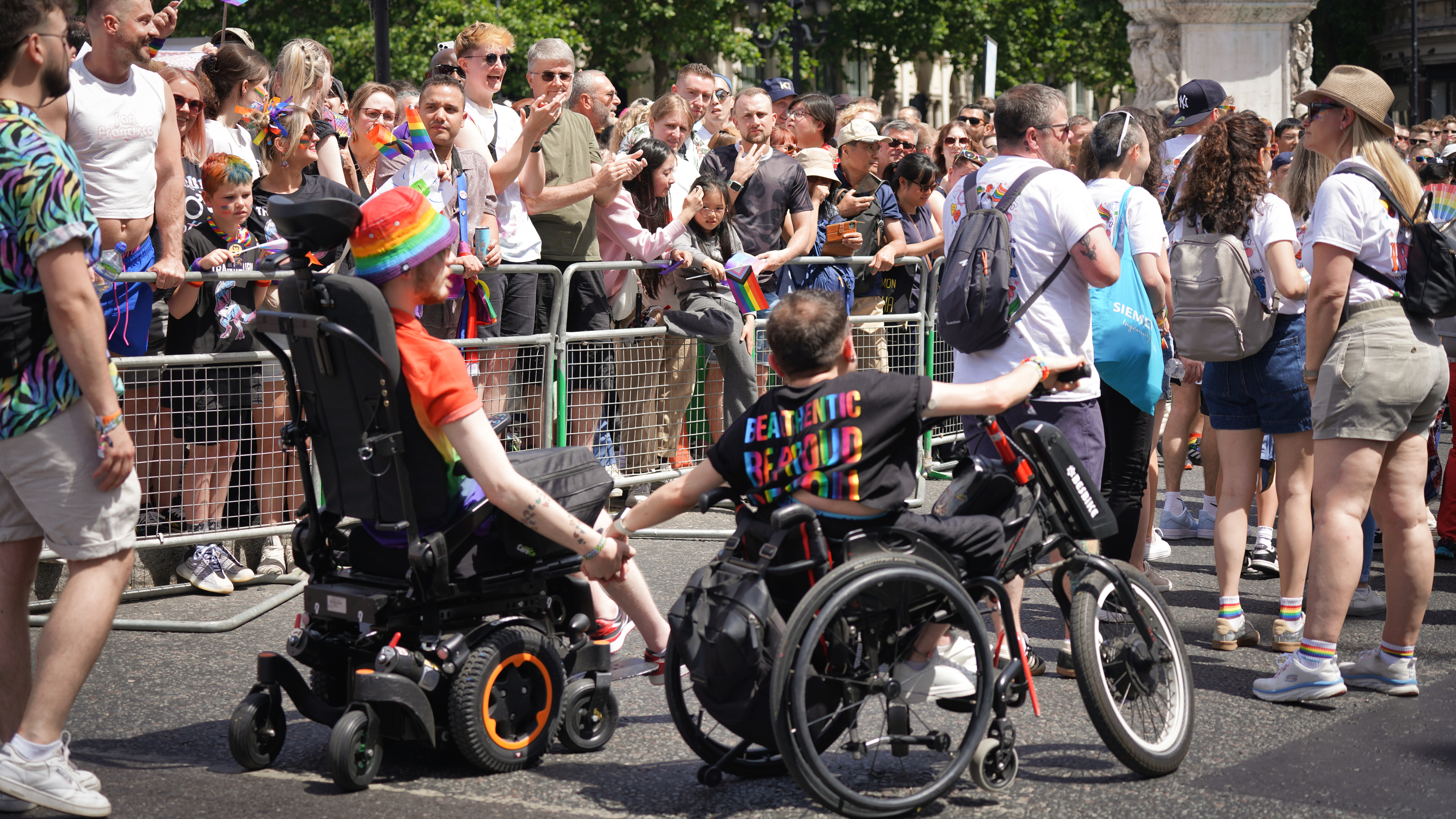 Two people in wheelchairs, one in a power chair and the other manual, hold hands while taking part in a Pride parade. They are surrounded by a cheering crowd, and one person wears a rainbow hat while the other’s shirt reads “BE AUTHENTIC BE PROUD.”