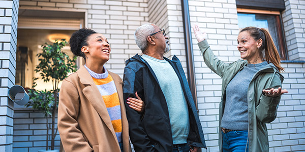 A young Black woman stands smiling arm-in-arm with an older Black man as they talk to a smiling white woman in front of a home