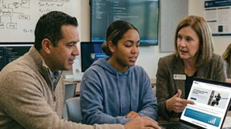 A female administrator points to a BICSI 529 Plan webpage on a laptop while consulting with a male parent and a female student in an ICT classroom.
