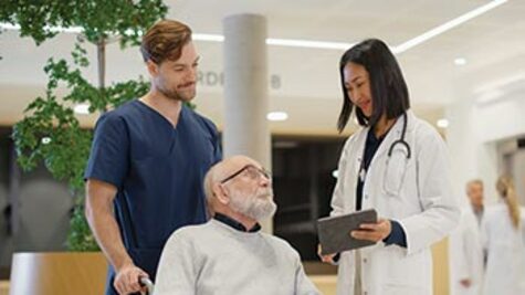 An elderly man in a wheelchair talks with a female doctor holding a tablet and a male nurse in a bright hospital lobby.