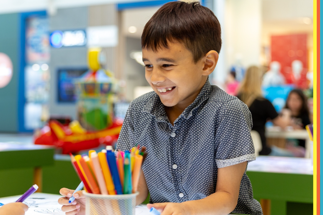 Smiling young child drawing with coloured pencils at a table in a shopping centre play area, with a blurred family-friendly background and colourful pencils in the foreground