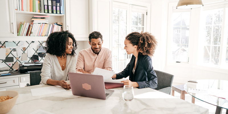 A couple goes over documents with a woman dressed in a suite in front of a laptop