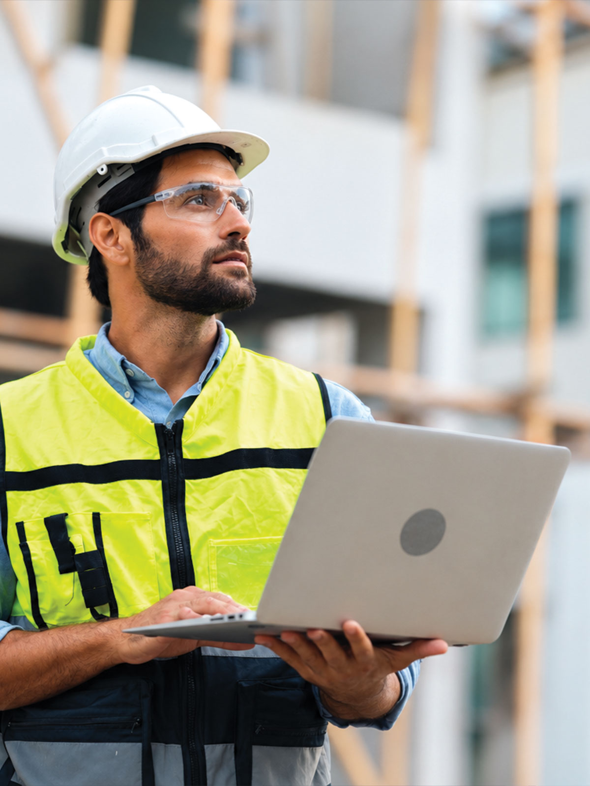 Field supervisor in a white hard hat and safety vest holding a laptop on an outdoor construction site