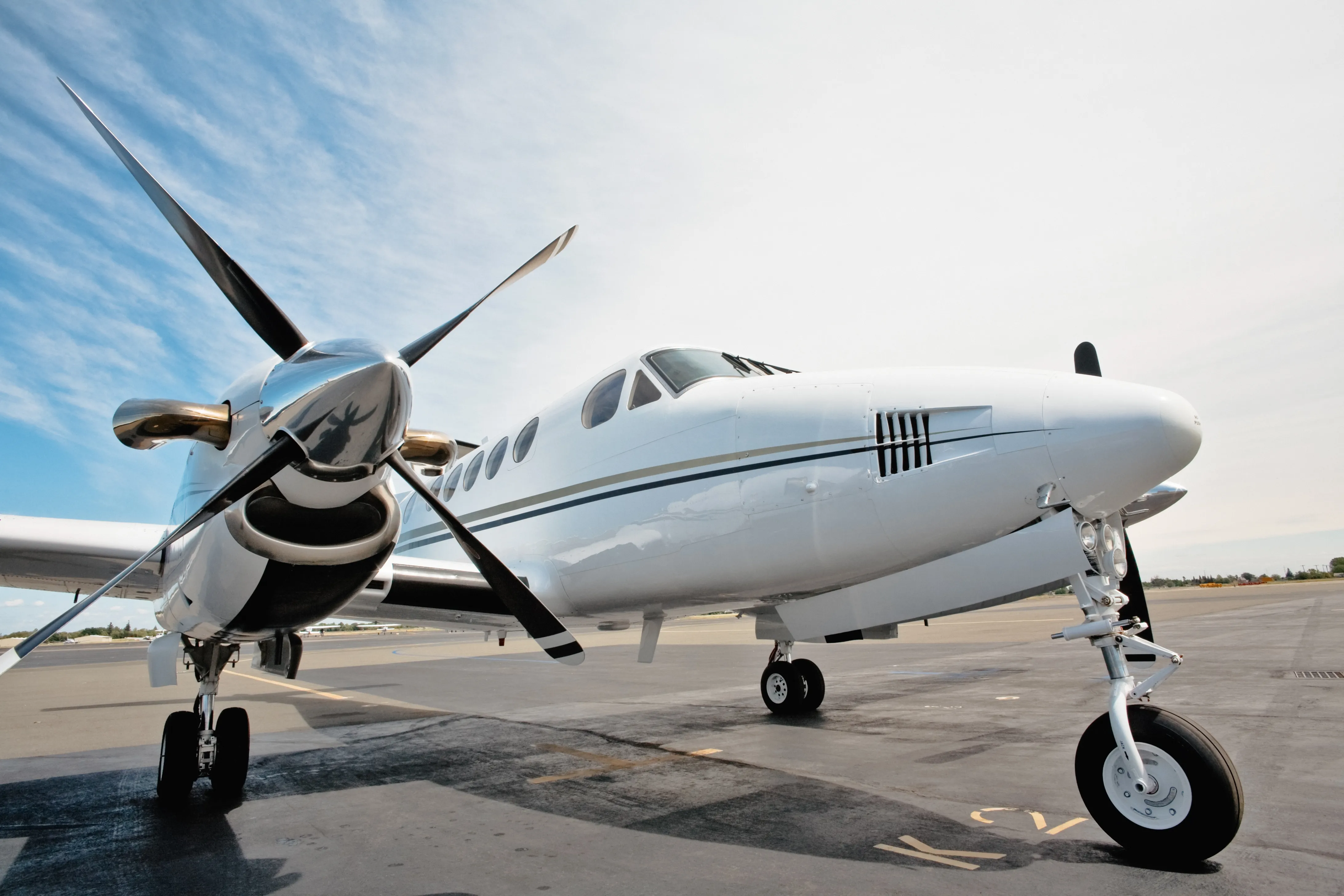 White twin engine airplane sitting on the tarmac. One twin engine is in the foreground, close to the camera. Blue sky with mid-level clouds in the background. 