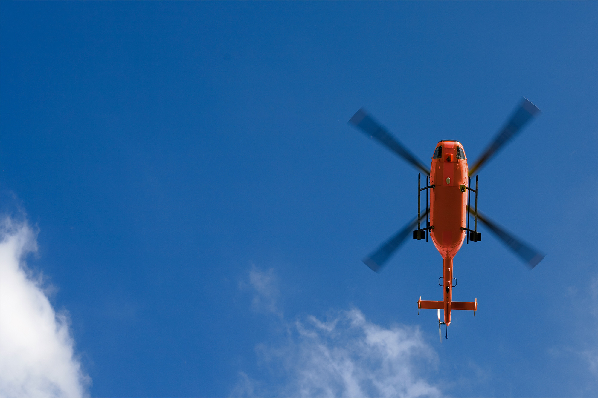 Photo of a red helicopter from CHC's fleet. Flying in the sky. Taken from below you can see the bottom of the helicopter as it flies over.