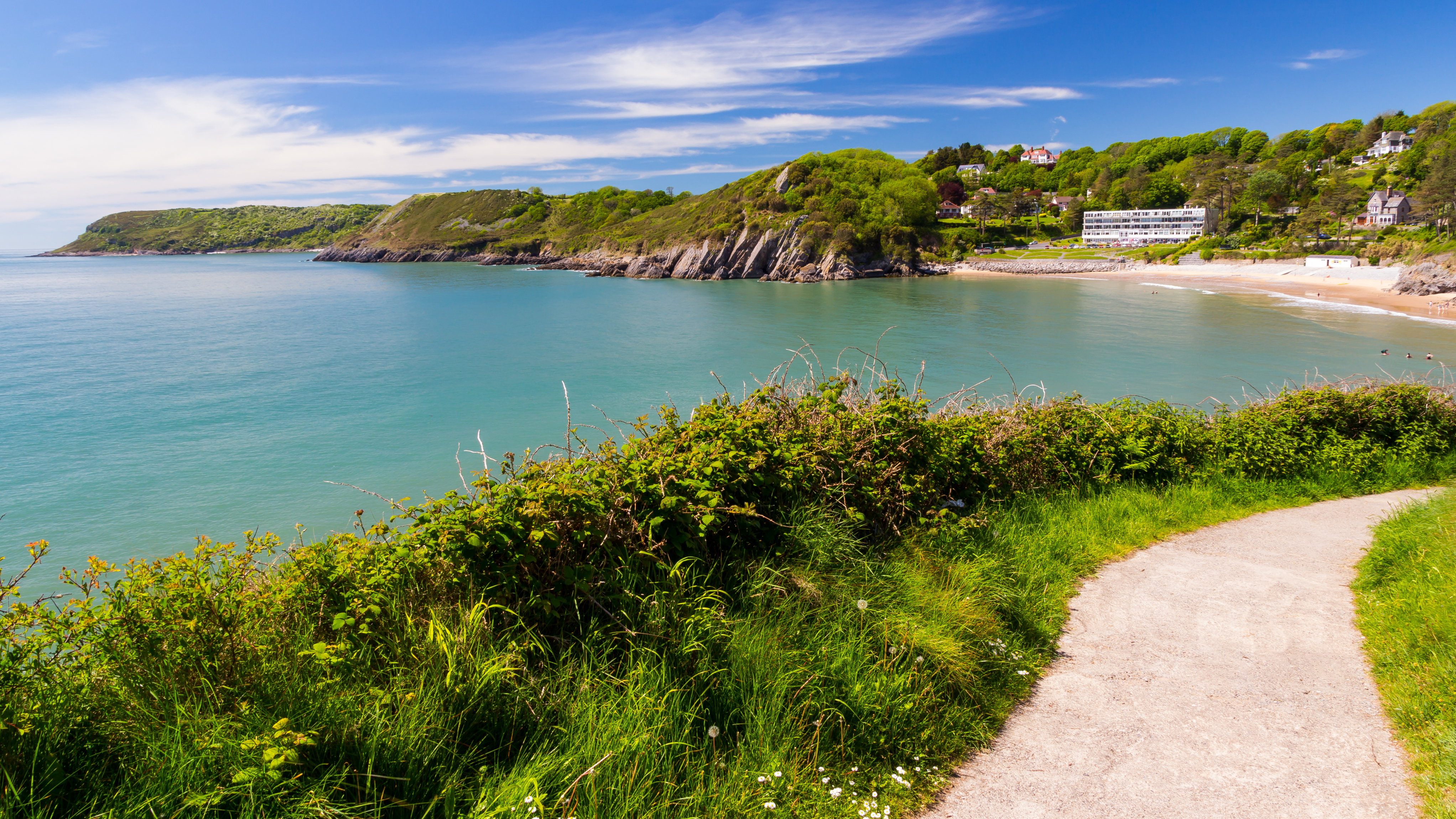 Caswell Bay on a sunny day, showing turquoise waters, tree-covered cliffs, and a sandy beach. A narrow path curves along the foreground, with lush greenery framing the coastal view.