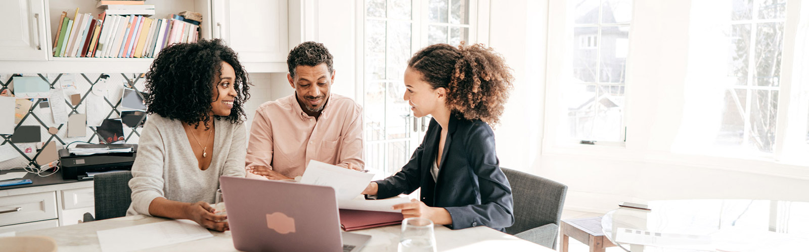 A Black man and a Black woman sit at a table covered with paperwork looking at a laptop screen with a Black woman wearing a suit jacket