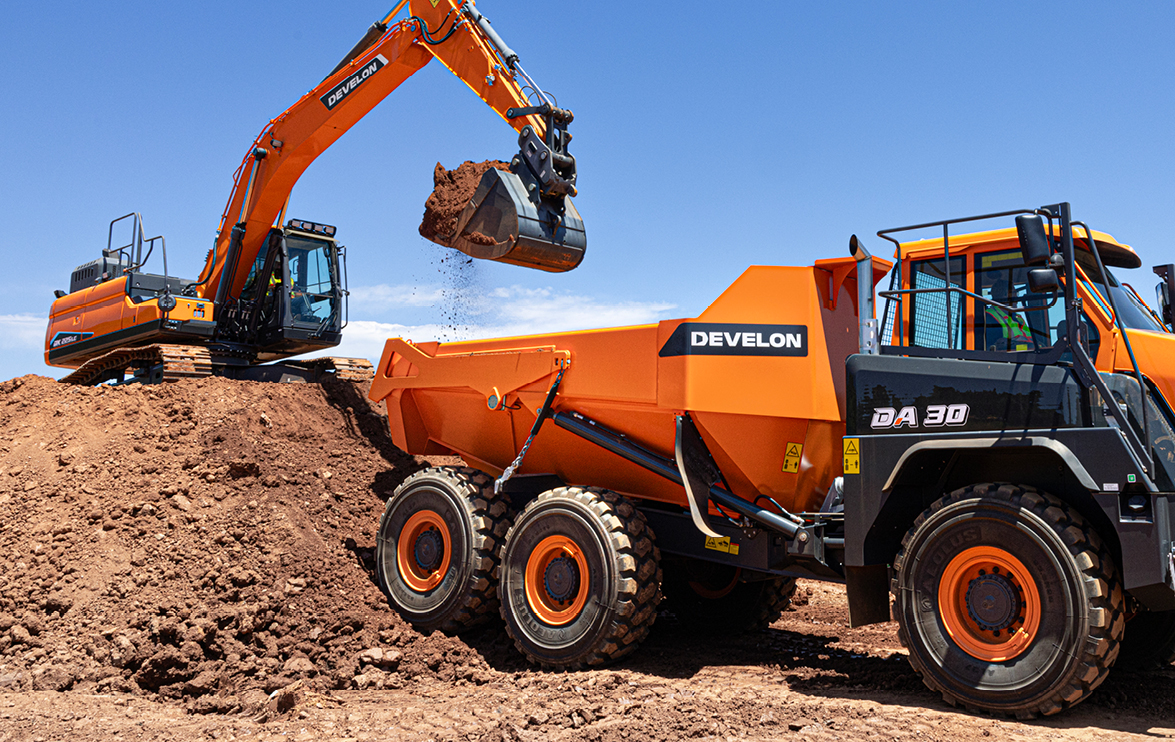 A DEVELON crawler excavator working on a large dirt mound dumps dirt into the bed of a DEVELON articulated dump truck.