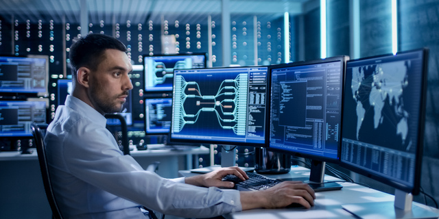 System Security Specialist Working at System Control Center. Room is Full of Screens Displaying Various Information.