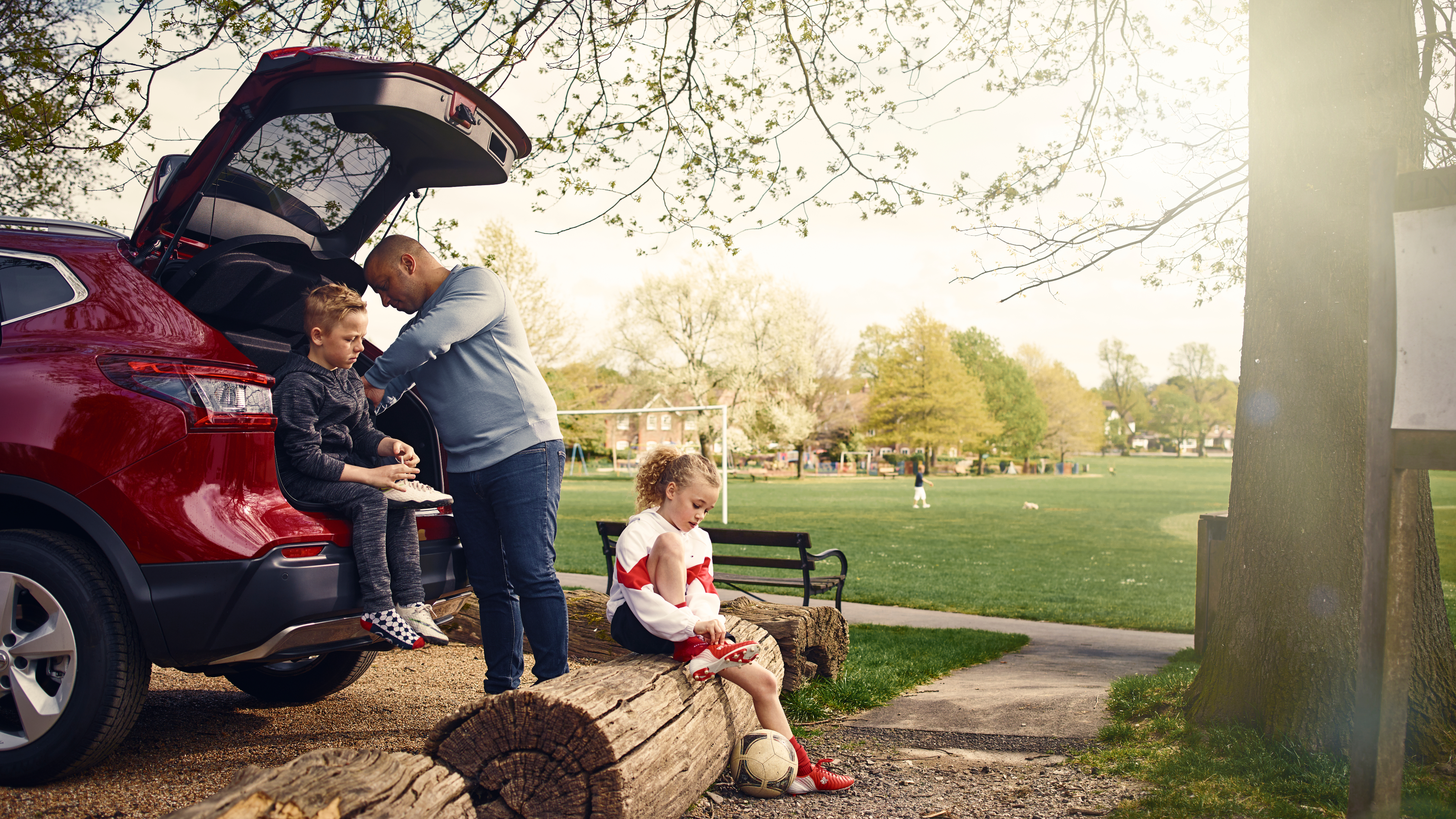 A man helps a young boy change shoes at the back of a red SUV parked beside a park, while a young girl in a football kit ties her laces on a nearby log with a ball at her feet. The park is filled with trees and people in the distance on a sunny day.