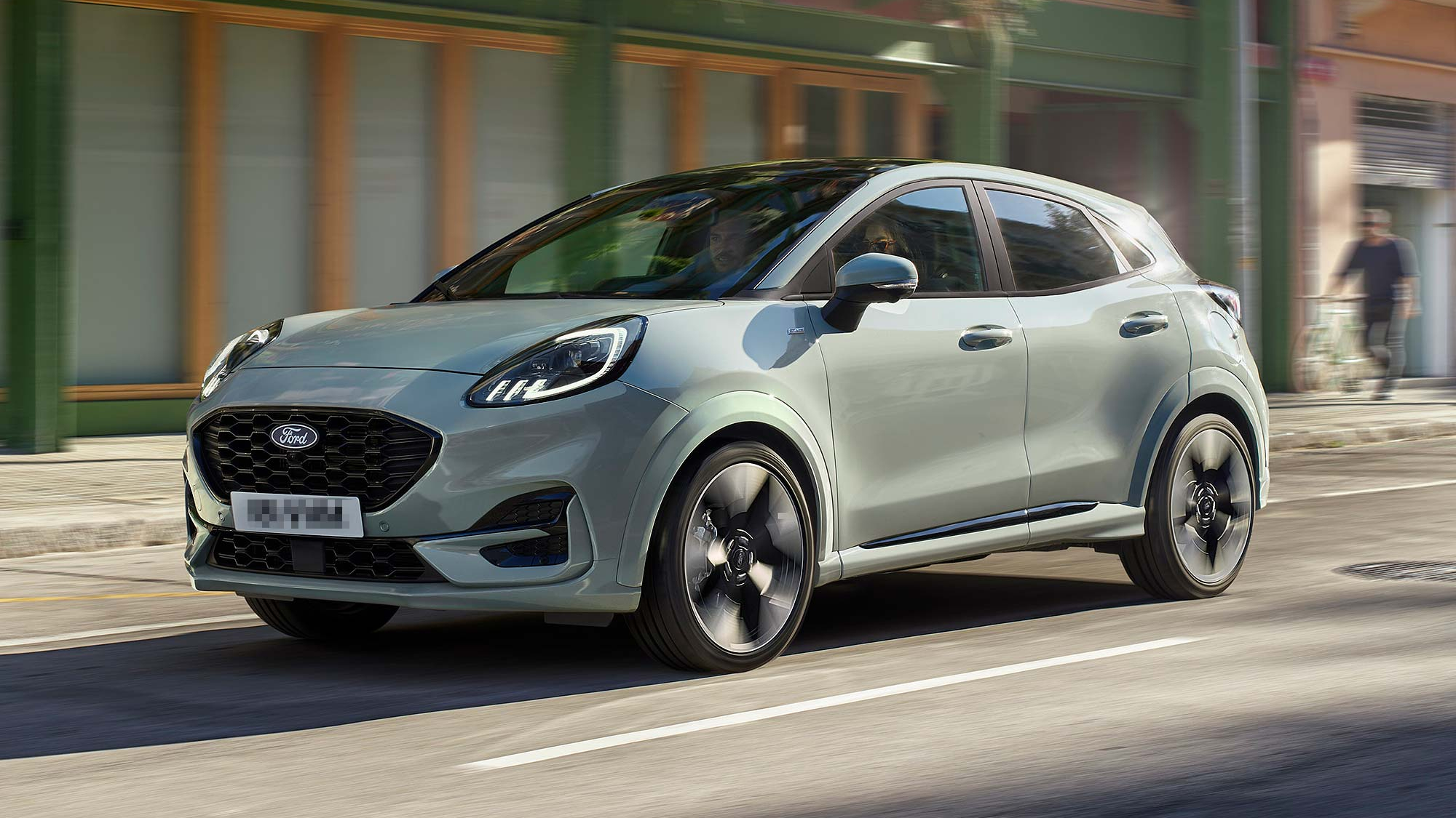 A grey Ford Puma compact SUV driving on a city street, featuring sleek headlights and a black mesh front grille.