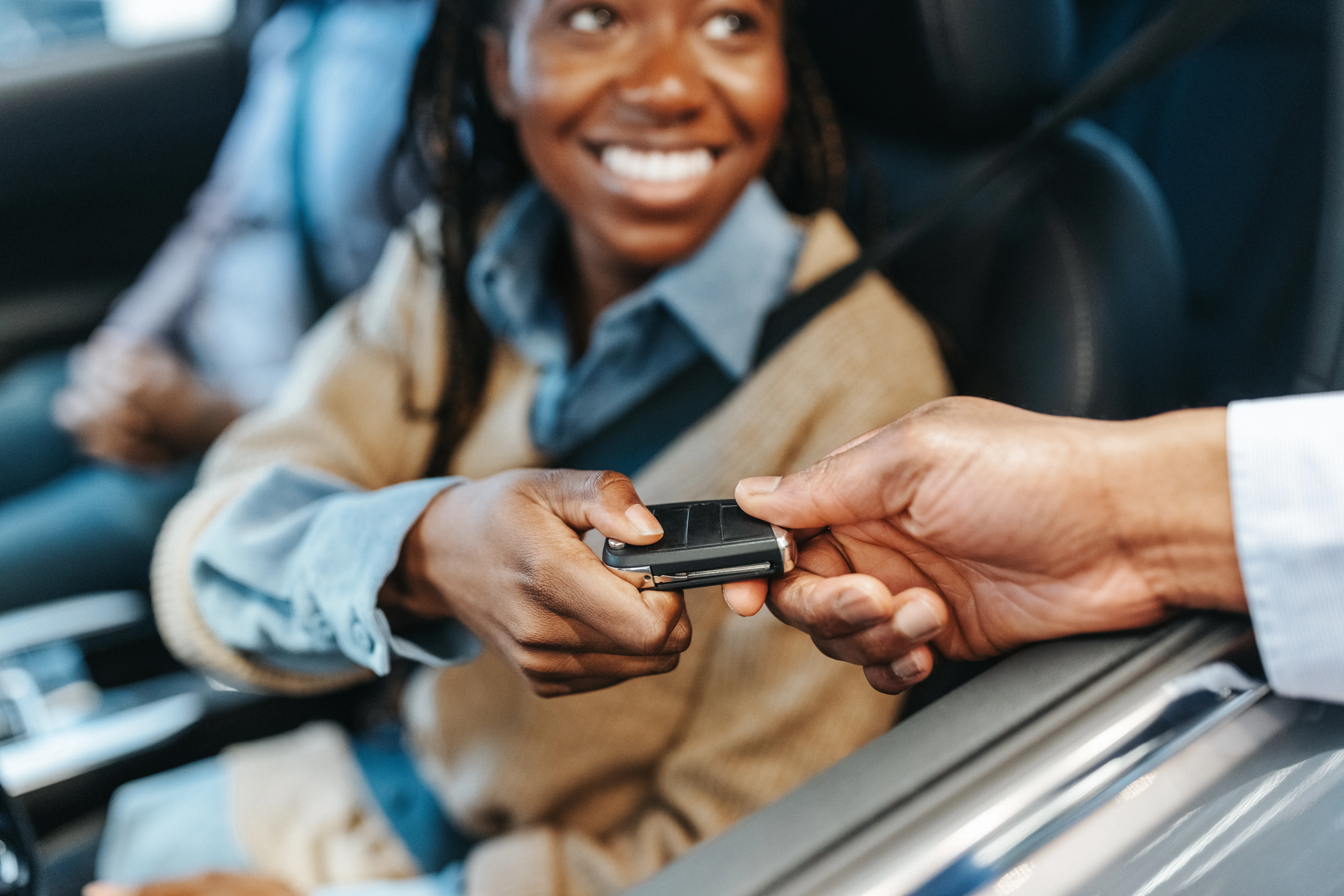 Close-up of salesman hand giving car keys to African American woman sitting inside car for test drive in the showroom.