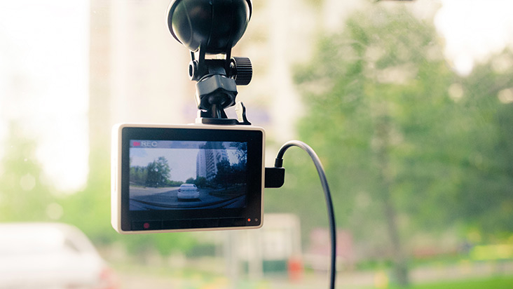 A dashcam mounted on a car windshield records the road behind, showing a white vehicle and buildings through the screen. The background is slightly blurred, with trees and urban scenery visible outside the window.