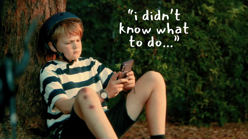 Young boy sits on the ground against a tree while looking at his phone, with the text 