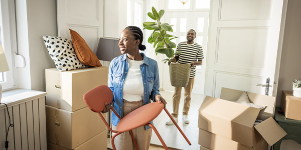A smiling Black woman holds a chair and a smiling Black man stand behind her holding a plant in their new home