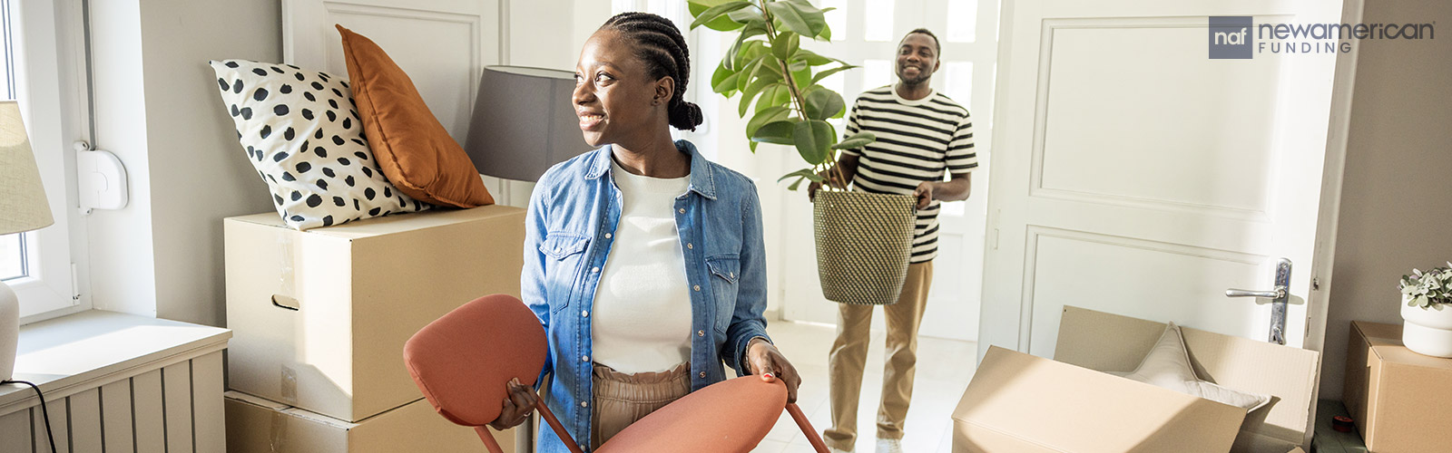 A smiling Black woman holds a chair and a smiling Black man stand behind her holding a plant in their new home