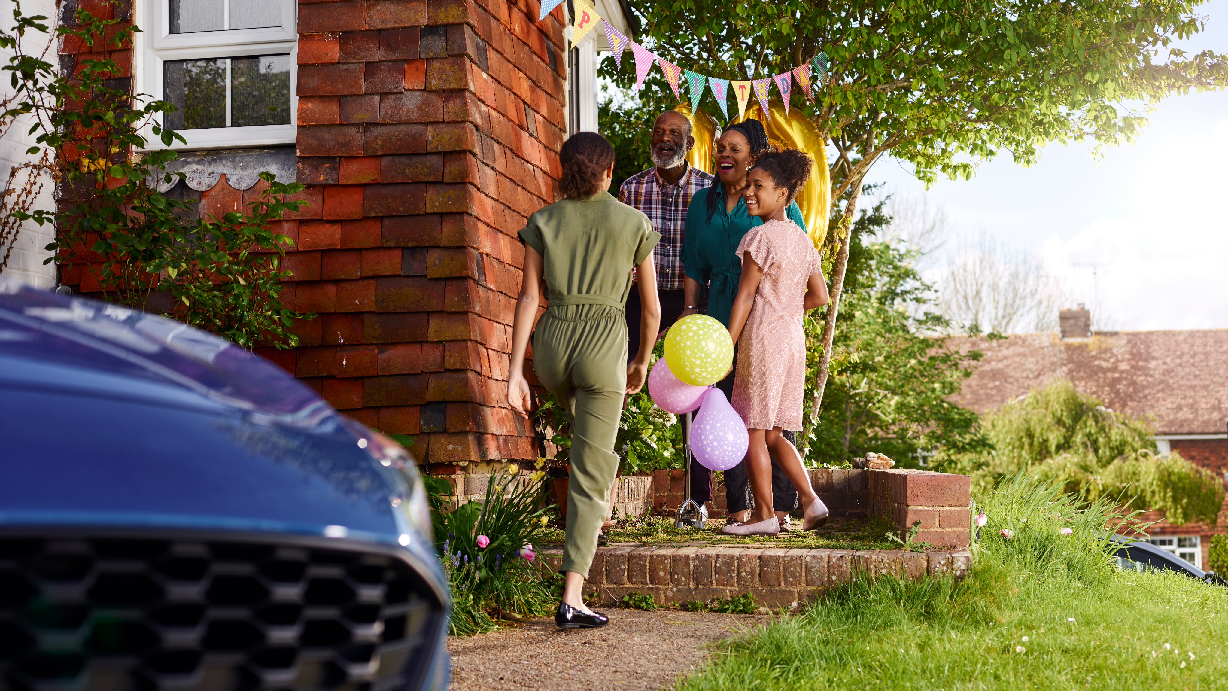 A girl walks up to a house holding balloons, greeted by smiling family members standing under a “Happy Birthday” banner outside the front door.
