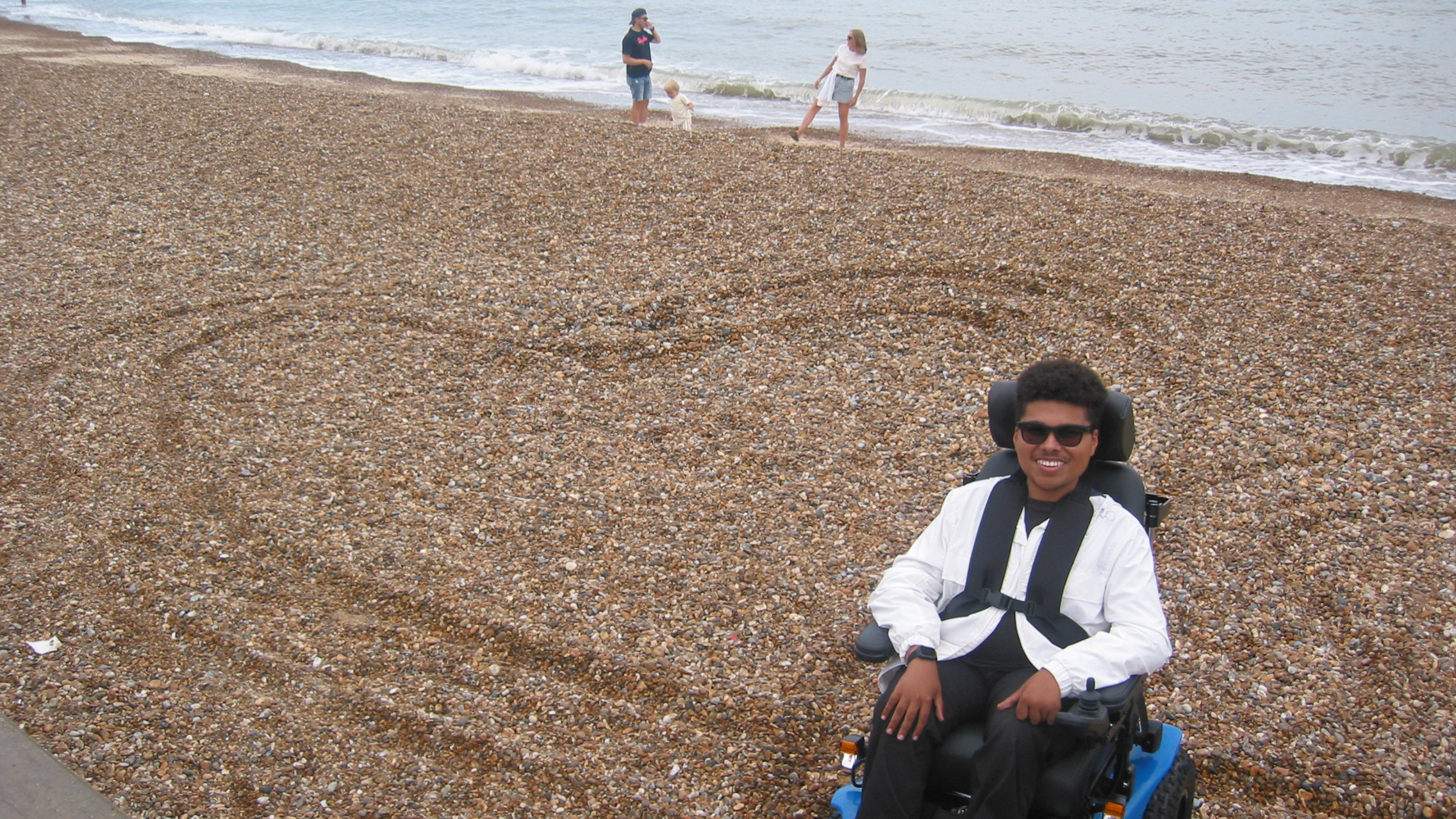 Maxwell McKnight sits in a powered wheelchair on a pebbled beach, smiling at the camera with a large heart shape drawn in the stones behind him.