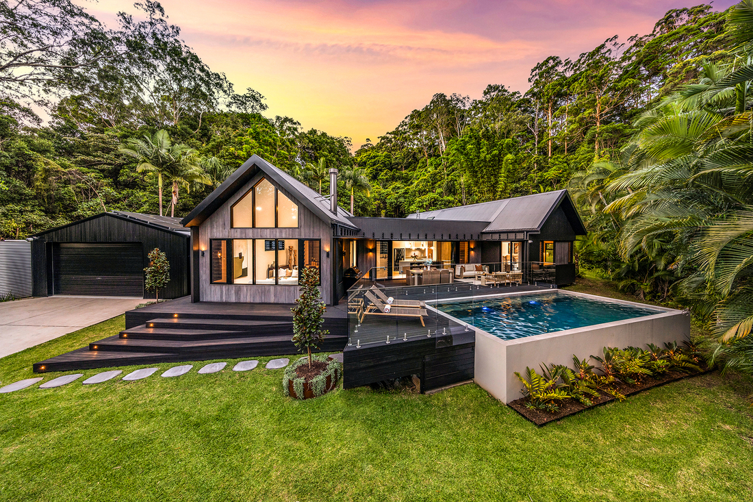 yourtown Prize Home Draw 545. Dusk external image from the front of a single-level pavilion-style house with black cladding. There is an alfresco area at the front with a pool. A garage and driveway is on the left hand side. Lush gardens surround the home.