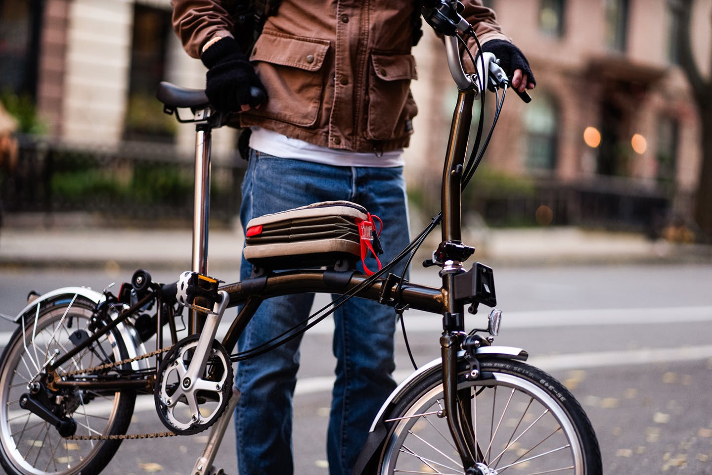 Closeup of a Brompton folding bicycle frame to which a collapsible brown fabric carrying case is affixed on the crossbar.