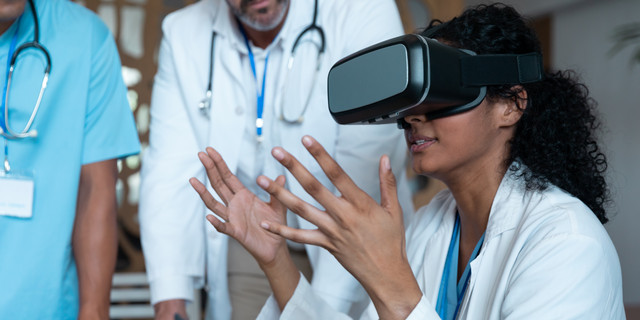 Diverse male and female doctors wearing face masks sitting at table and using vr glasses