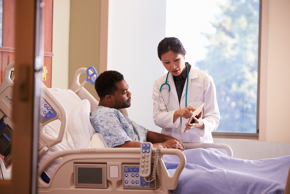 A doctor operates a handheld tablet while talking with a patient in a hospital bed.