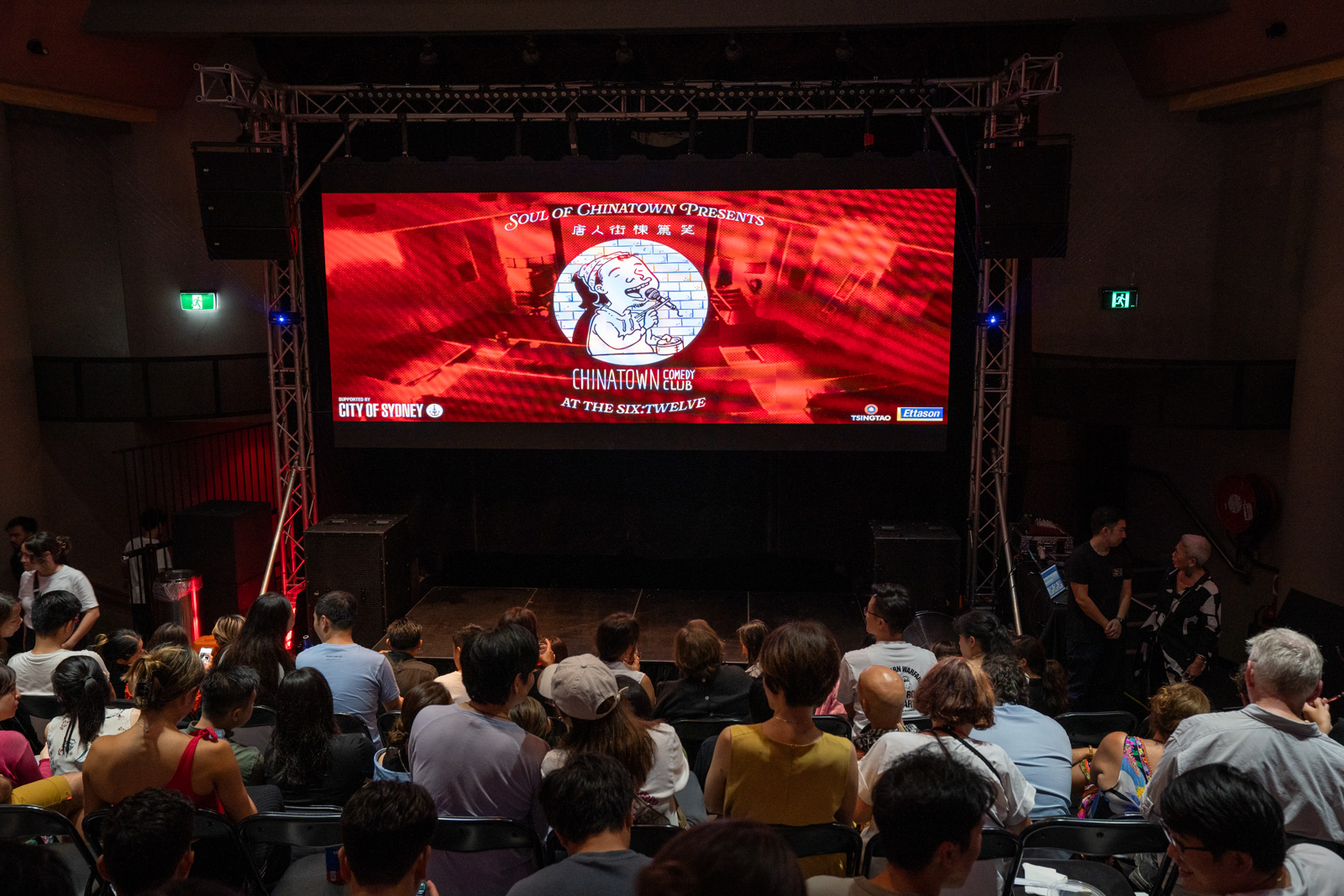 crowd seated in tiers with a stage at the front with 'Chinatown Comedy Club' on the screen