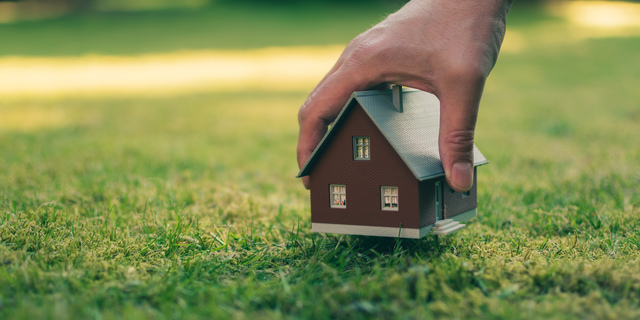Concept of selling a house. A hand is holding a model house above green meadow.