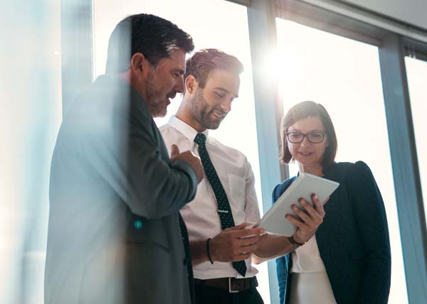 Three business professionals standing near a window, looking at a digital tablet together.