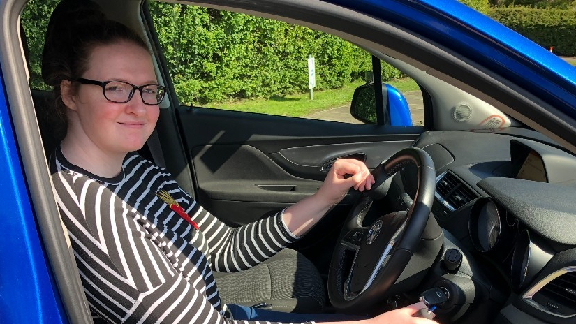 Shona Louise is wearing glasses and a black-and-white striped top smiles while sitting in the driver's seat of a blue car. She has one hand on the steering wheel and the other on the ignition, ready to drive on a sunny day.