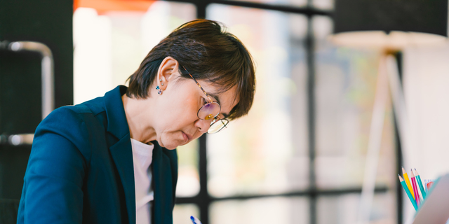 Beautiful middle age Asian woman working on paperwork in modern contemporary office, with laptop computer. Business owner, entrepreneur, executive manager, or employee office worker concept