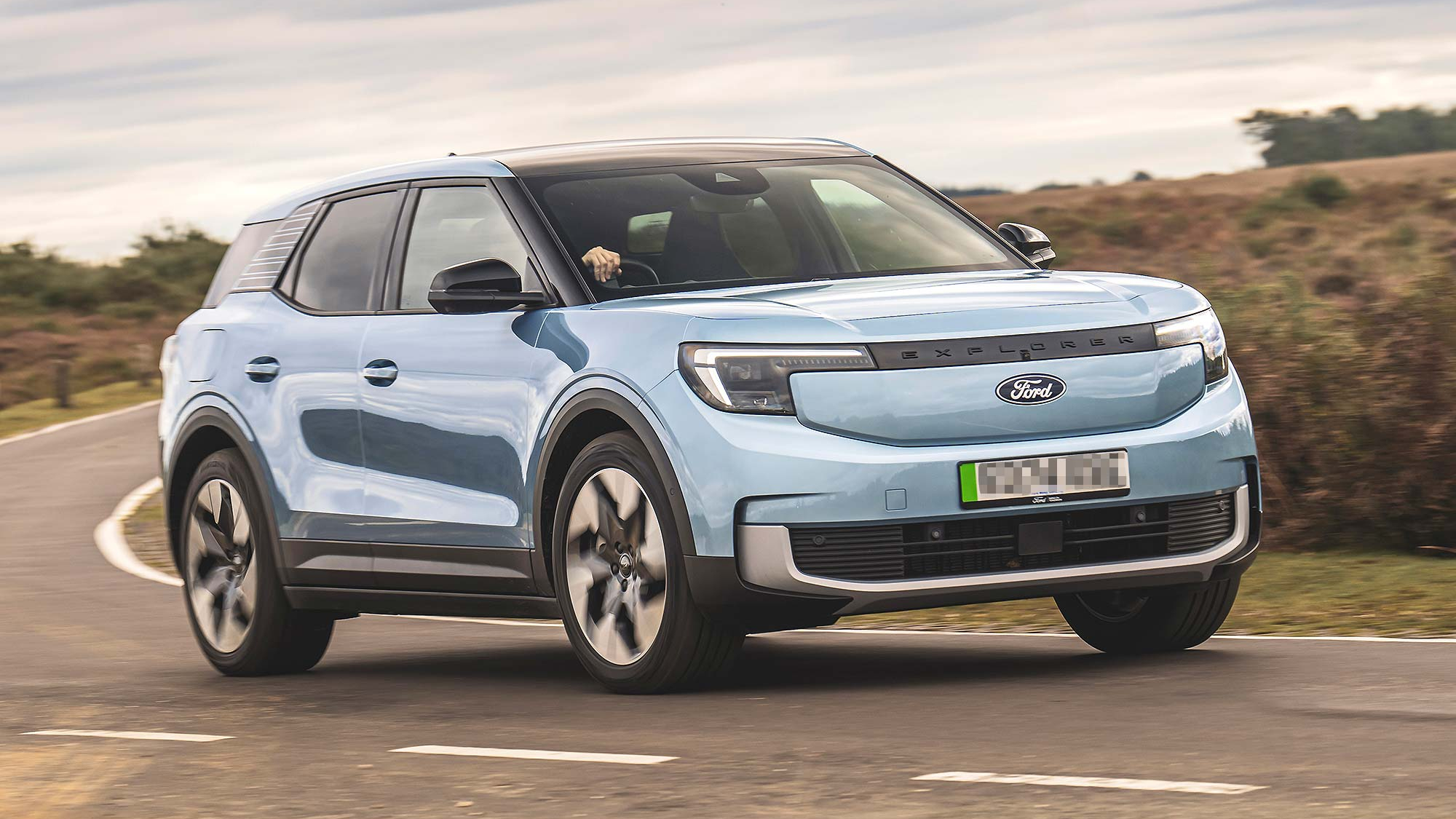 A light blue Ford Explorer EV driving on a winding country road, showing off its bold front and aerodynamic profile.