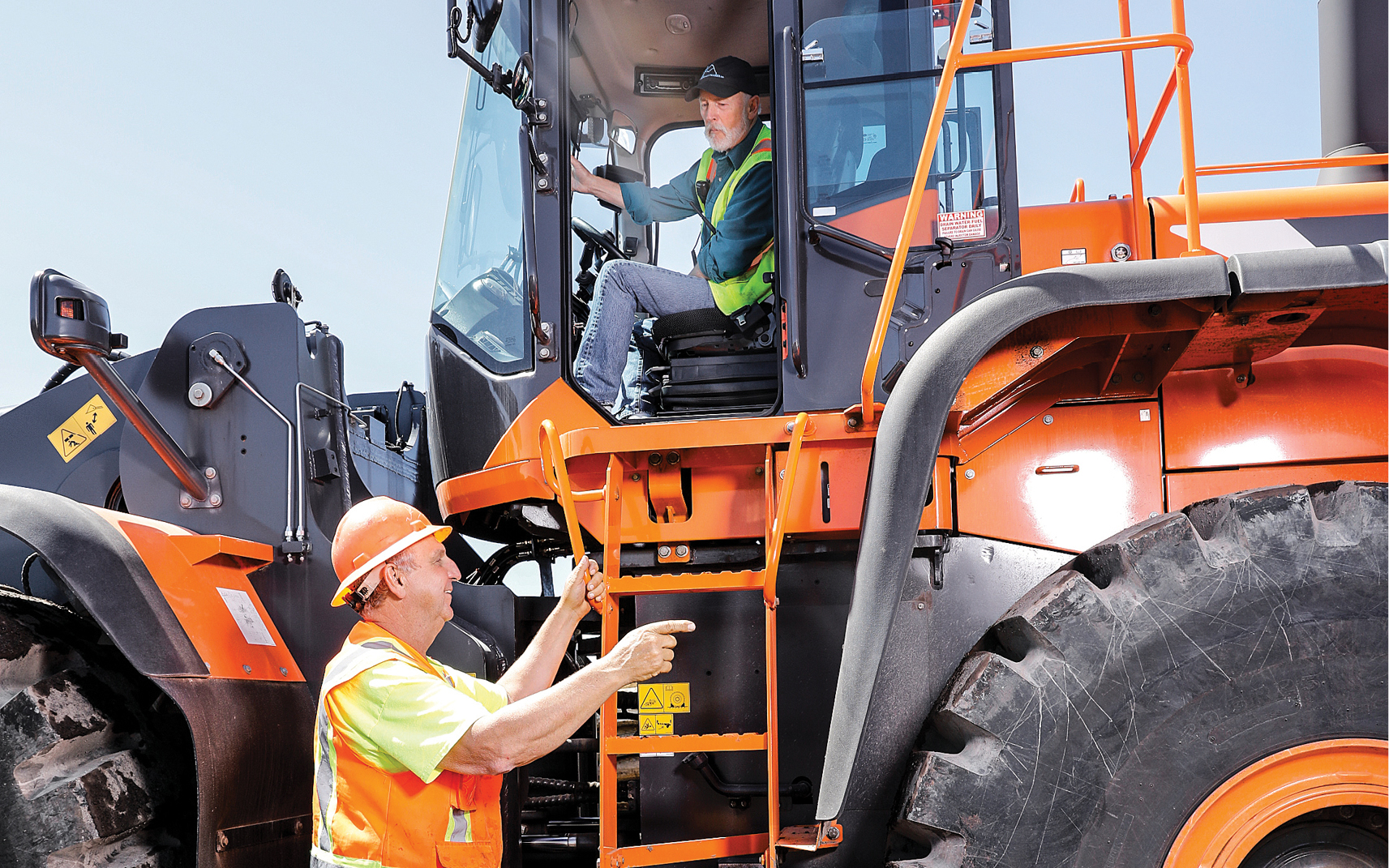 A wheel loader operator visits with a worker standing next to the machine.