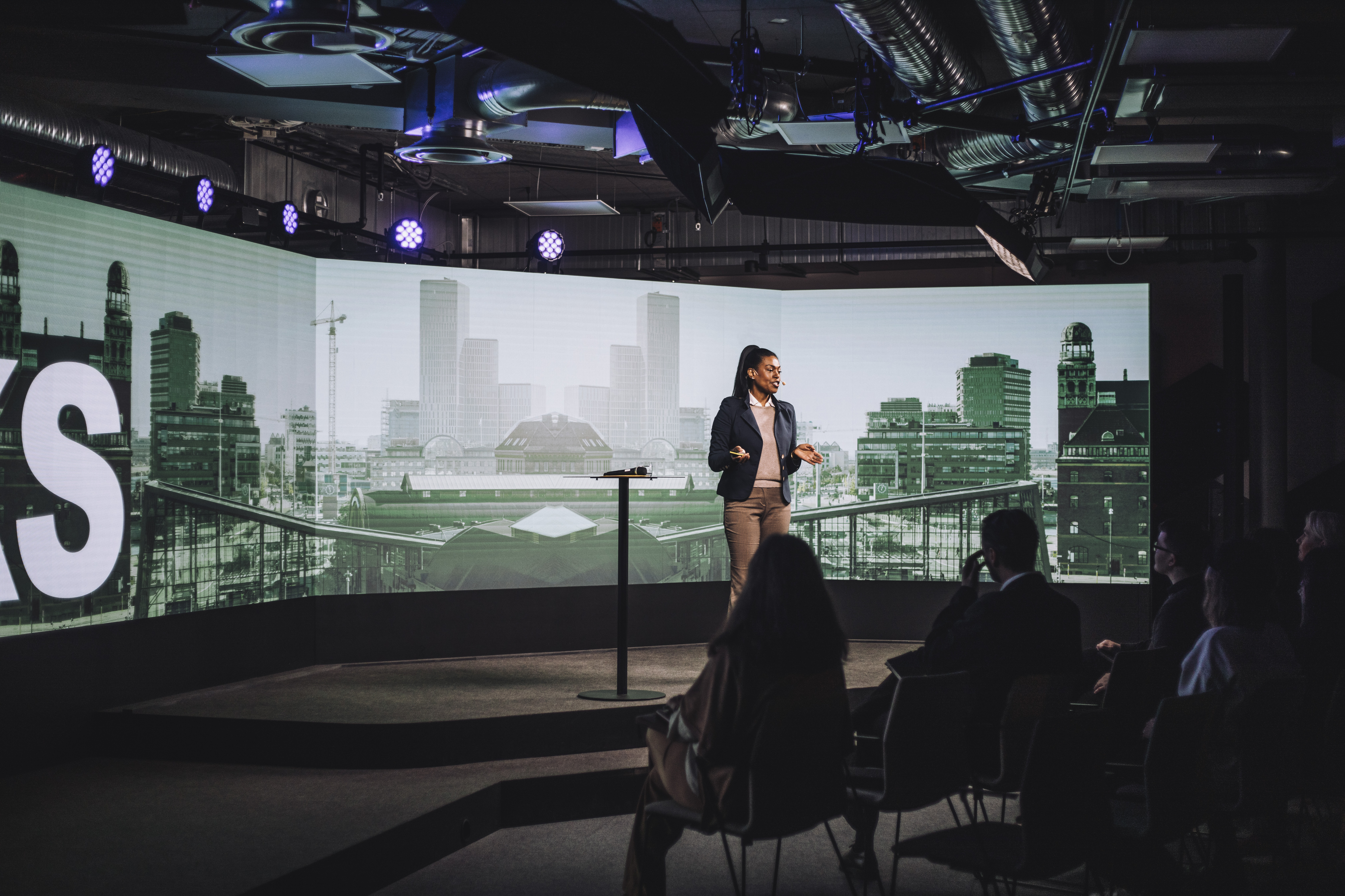 A woman stands in front of a 4k projector screen giving a presentation in a dark room to colleagues.