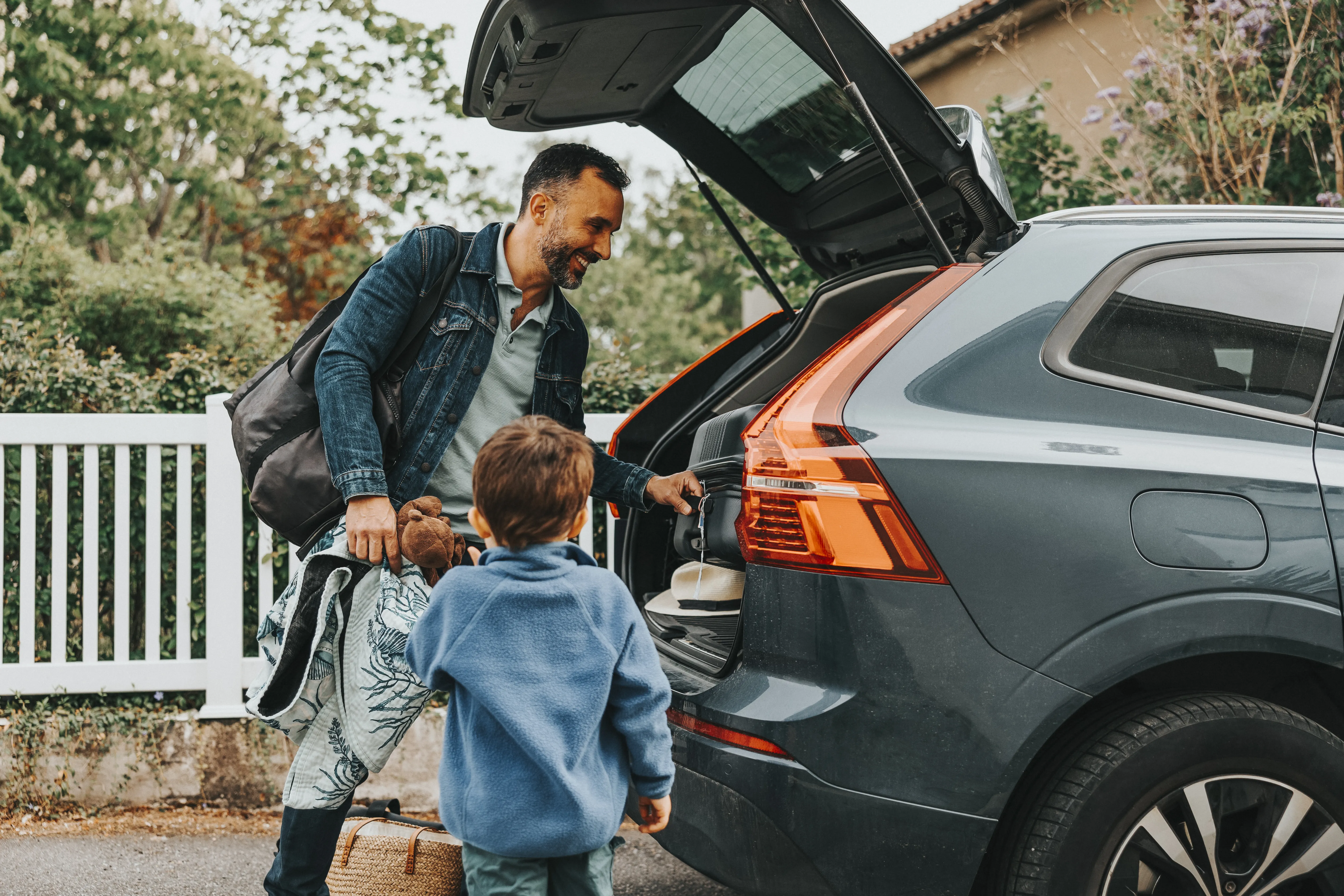 Smiling man loading luggage with son standing near car trunk. 