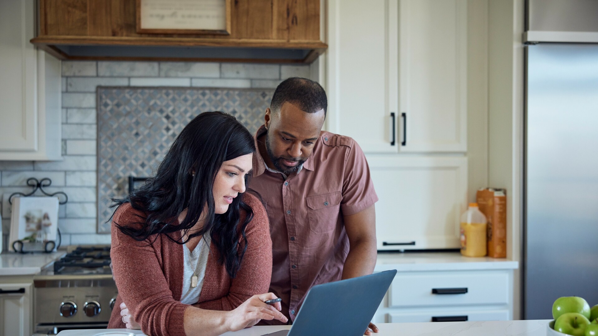 Caucasian woman and African American man sit at kitchen counter with breakfast working with pen, paper and laptop.