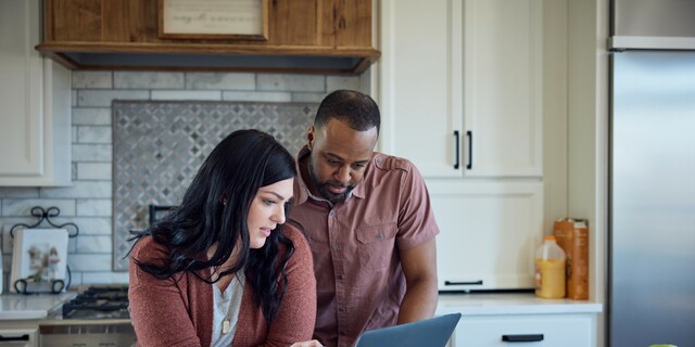 Caucasian woman and African American man sit at kitchen counter with breakfast working with pen, paper and laptop.