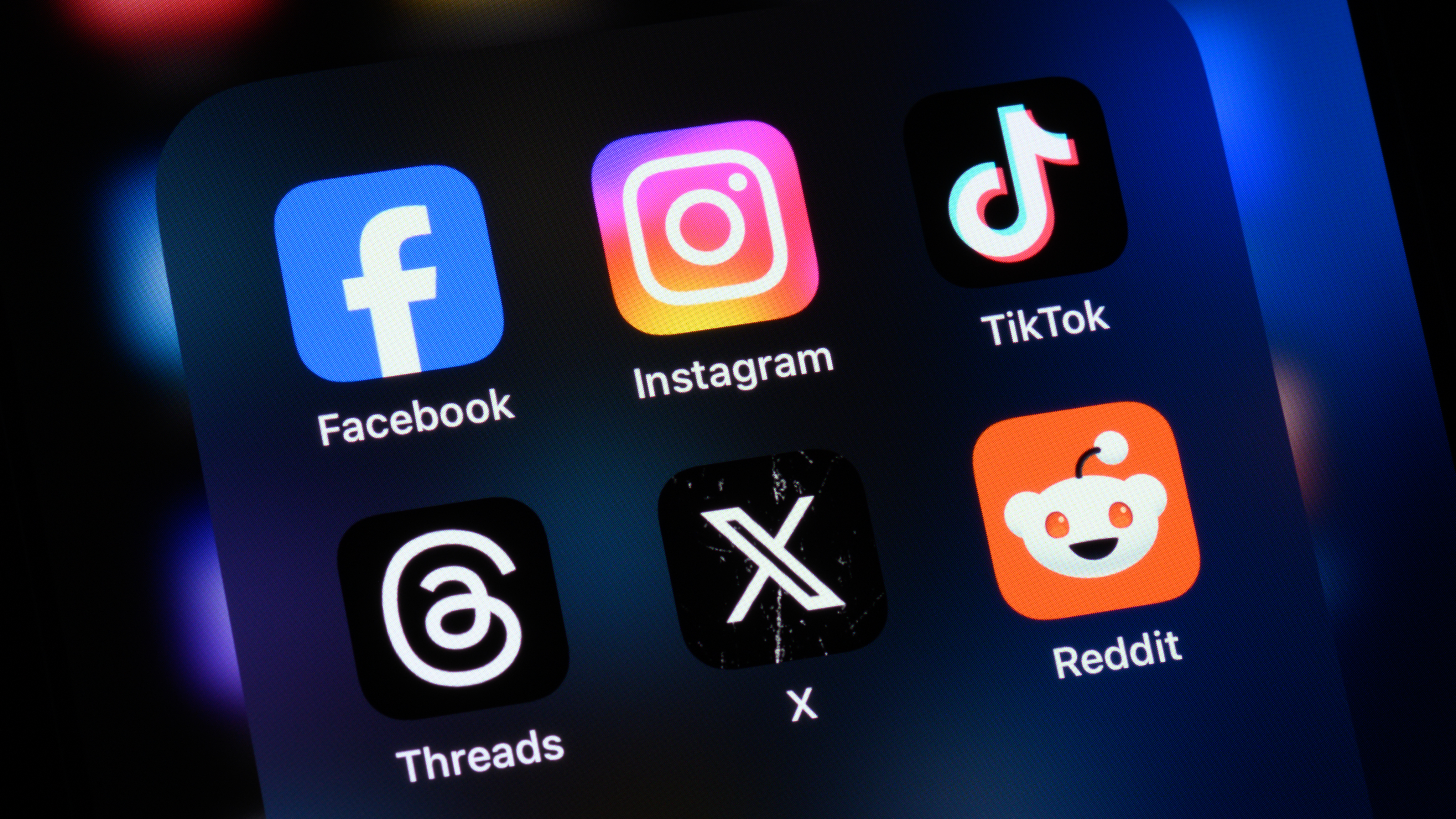 A close-up of a smartphone screen displaying six popular social media app icons: Facebook, Instagram, TikTok, Threads, X (formerly Twitter), and Reddit. Each icon is brightly colored and clearly labeled, standing out against a dark background with blurred lights, highlighting the phone's focus on social connectivity