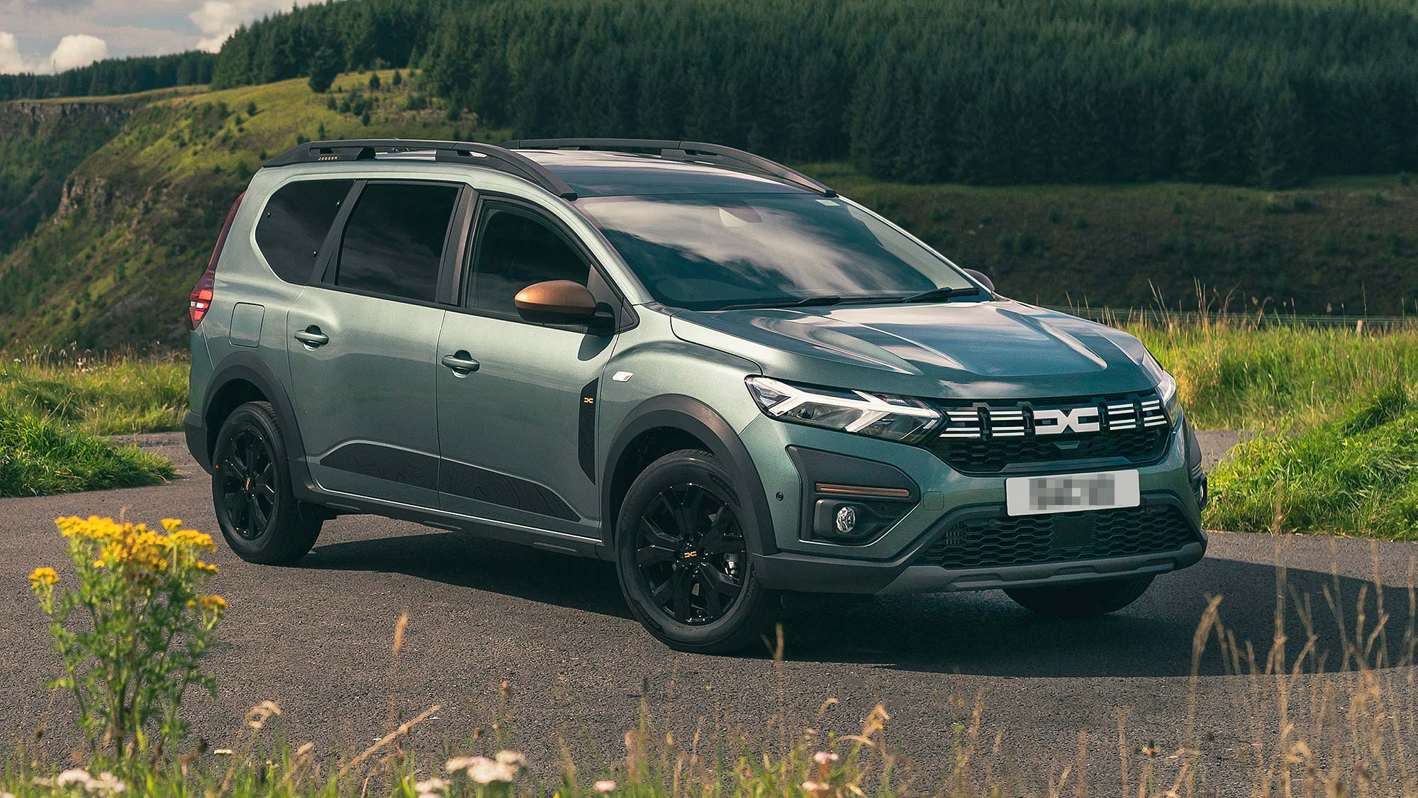 A green Dacia Jogger crossover SUV parked on a scenic countryside road, with roof rails and rugged styling.